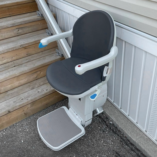 Outdoor stair lift with dark gray cushioned seat and armrests installed along a straight rail.