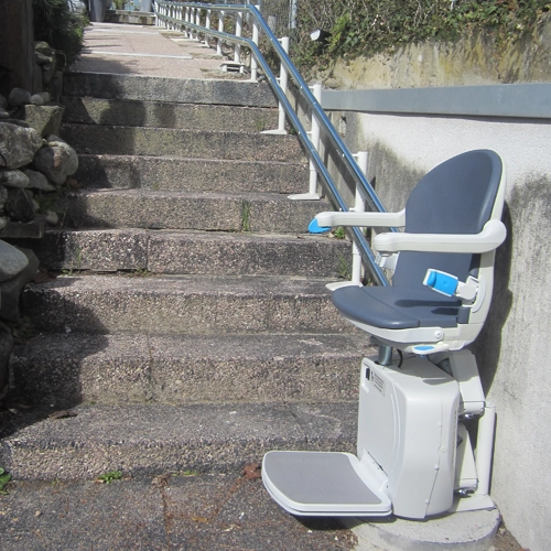 Curved stair lift attached to a rail along a stone staircase in an outdoor setting.
