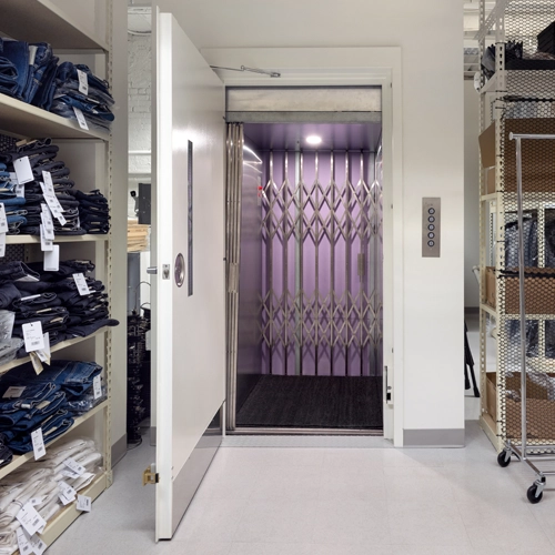 Retail‑style material lift with a white swing door open to reveal a cab with a purple accordion gate, installed between shelving units and storage racks in a commercial back‑of‑house area.