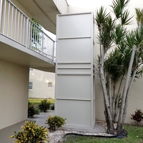 Outdoor vertical platform lift enclosed in a light-colored shaft next to a building walkway.