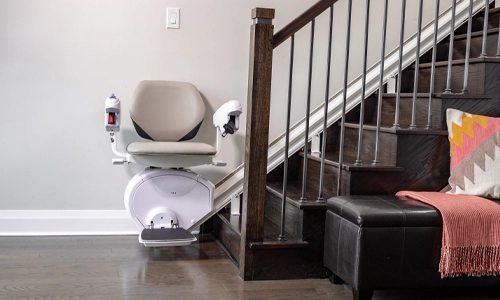 Stairlift parked at the bottom of a wooden staircase, featuring a padded seat, armrests, and footrest mounted on a rail along the side of the stairs in a modern home interior.