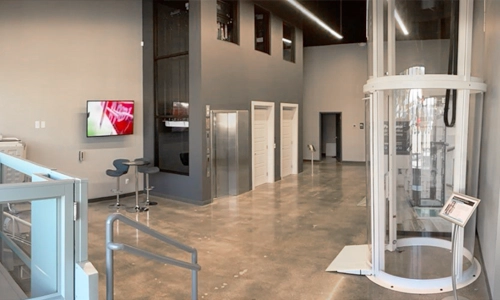 Showroom in Maryland featuring a glossy concrete floor, modern elevator displays, a cylindrical glass lift on the right, wall‑mounted screens, sample doors along the back wall, and contemporary seating near the entrance railings.