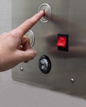 Hand pressing a metal elevator control panel with illuminated buttons and a keyed switch.