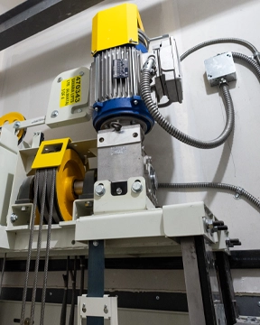 LULA Elevator drive system with a motor, gearbox, pulleys, and steel cables mounted on a structural frame, showing electrical conduit and control components along the wall.