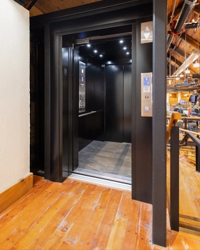 Modern elevator with a matte black frame and open cab, featuring dark interior panels, recessed ceiling lights, and wood‑tone flooring, set within a space with exposed beams and warm wooden floors.