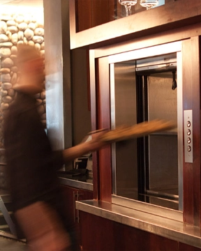 Blurred person placing a long food tray into an open stainless steel material lift built into a wood‑framed counter.