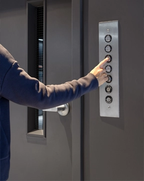 Hand pressing a control button on a wall-mounted panel beside a closed vertical material lift door.