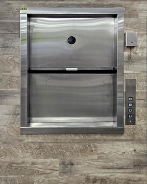 Stainless steel material lift with a closed vertical sliding door and control panel mounted on the right side.