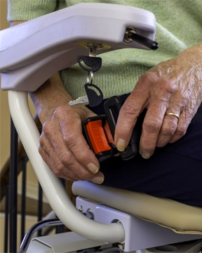 Hands fastening a stairlift seatbelt on a padded chair with an armrest and key switch above.