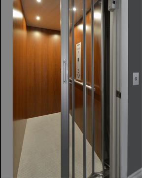 Interior view of a home elevator with wood-paneled walls, recessed ceiling lights, a metal accordion gate, and a light-colored floor.