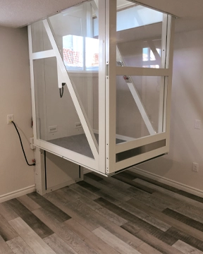 Through‑the‑floor lift with a white framed enclosure shown raised between levels, featuring clear panels and positioned above a laminate floor.