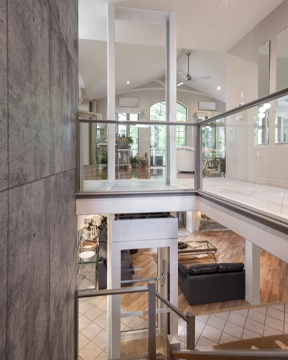 Through‑the‑floor lift installed in a multi‑level home, shown traveling between an upper landing with glass railings and a lower living area.