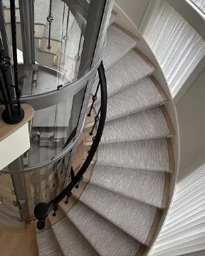 Glass home elevator centered within a curved staircase with light carpeted steps and a black handrail.