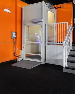 Indoor vertical platform lift with a clear door and white frame, installed beside a short staircase with white railings against an orange wall.
