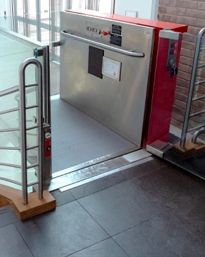 Stainless steel inclined platform lift at the base of a staircase, shown with its platform raised and side safety panels in place, positioned beside glass and metal railings.