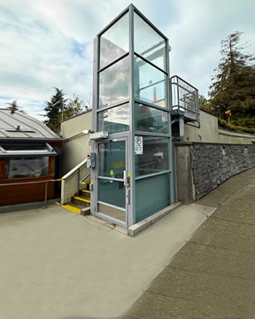 Outdoor vertical platform lift with a clear glass enclosure and metal framing, positioned beside a ramp and stone retaining wall, providing accessible travel between levels.