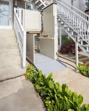 Outdoor vertical platform lift installed beside a concrete staircase, shown with an extended ramp, safety panels, and surrounding garden plants along the walkway.