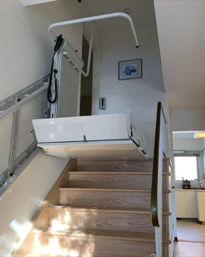 Inclined platform lift positioned on a wooden staircase, shown with the platform folded upright and mounted rail running along the wall.