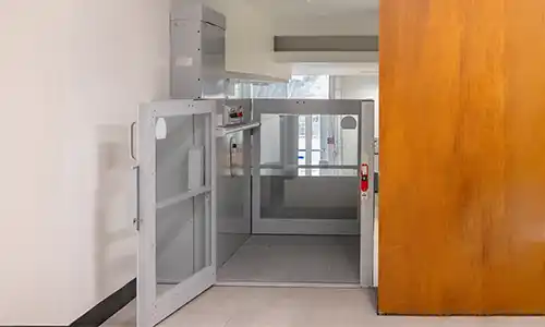 Indoor vertical platform lift with glass enclosure and swing gate installed in a commercial or residential hallway, providing wheelchair‑accessible access between levels.