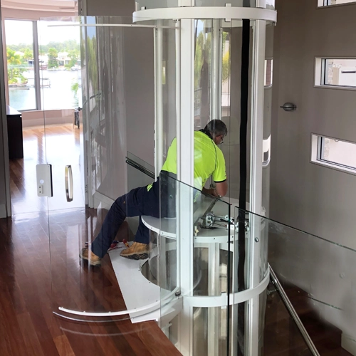Elevator technician servicing a panoramic glass residential elevator, performing maintenance inside a cylindrical home lift in a luxury multi‑level interior.