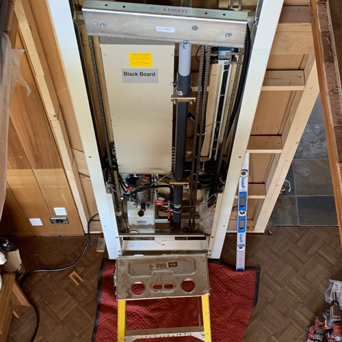 Residential home elevator machinery exposed inside an unfinished shaft, showing motor assembly, guide rails, wiring, and mechanical components during installation or service.