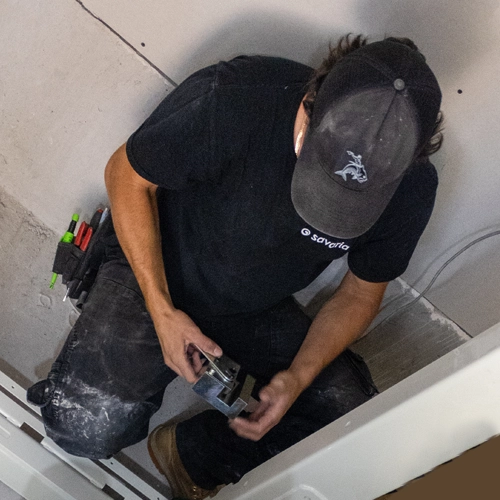 Elevator technician installing hardware components inside a residential home elevator shaft, kneeling on the floor with tools while completing lift installation work.