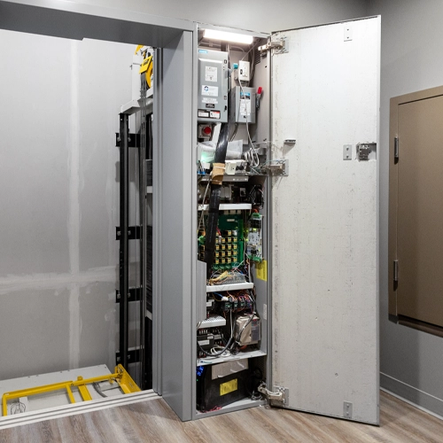 Residential home elevator with open control cabinet door, exposing electrical controller, wiring, circuit boards, and lift components installed in a finished interior hallway.