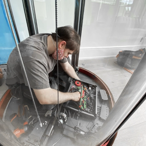 Elevator technician wearing a face mask performing maintenance on electrical components inside a panoramic glass residential elevator cabin.