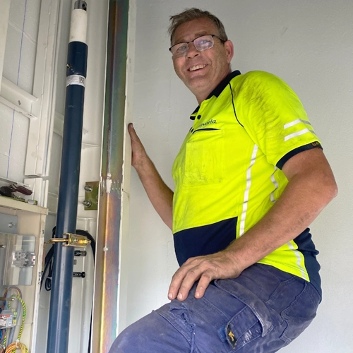 Smiling elevator technician wearing high‑visibility safety shirt, standing beside residential elevator piping and mechanical components during lift installation or service.