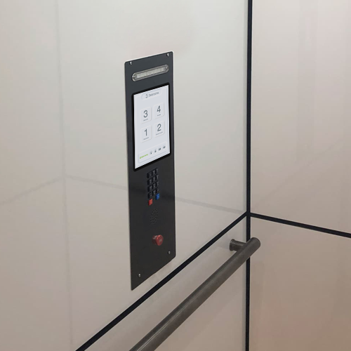 Blackened stainless steel elevator control panel with touchscreen floor buttons, emergency stop, speaker grille, and handrail inside a modern residential lift cabin.