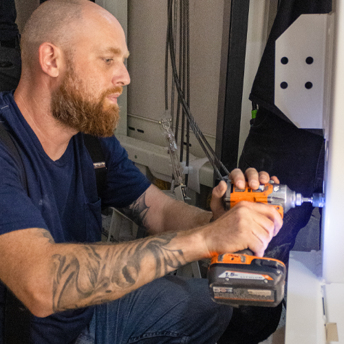 Elevator technician performing maintenance using a power drill inside a residential lift shaft, installing and servicing home elevator components.