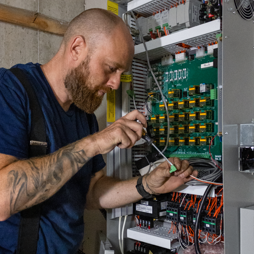 Elevator technician servicing a residential elevator control panel, adjusting wiring and electrical components inside a lift controller cabinet.